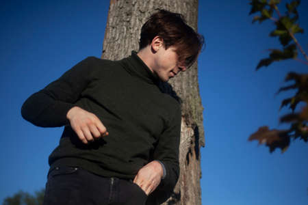 The guy is standing by a tree. A young man in a half-dress. A man in the woods. Outdoor recreation. The guy poses during the day.の写真素材
