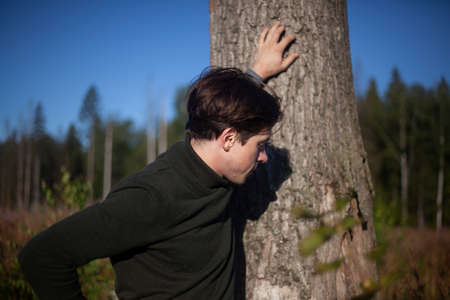 The guy is standing by a tree. A young man in a half-dress. A man in the woods. Outdoor recreation. The guy poses during the day.の写真素材