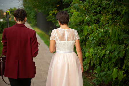 Two girls are walking down the street in different clothes. A girl in a dress and her friend in a formal jacket. The bride walks around the city.の写真素材