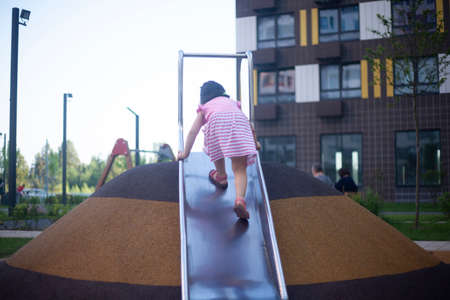 The girl climbs the slide in the playground. The child plays in the street. A girl with black hair runs along a metal slide.の写真素材