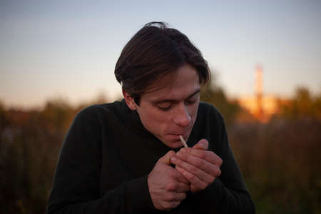 The guy smokes a cigarette. A man with a cigarette in the morning. A young man on a morning walk. Harm to health from tobacco. The young man rolled a roll of marijuana.の写真素材