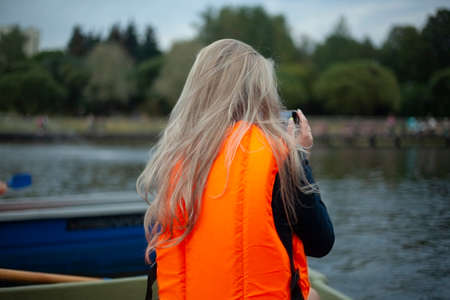 Life jacket on the girl. Orange life jacket on a woman sailing in a boat. Girl with phones in hands on the water.の写真素材