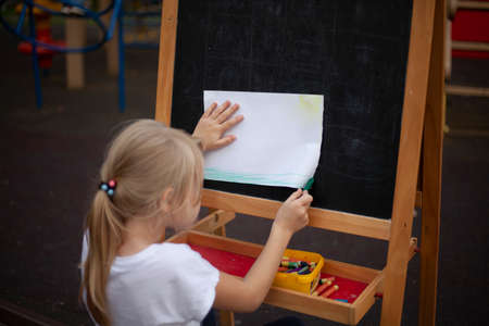 The child draws at the easel on the street. Painting lessons in the open air. A young artist is painting a picture. A girl in a white T-shirt sits with her back.の写真素材