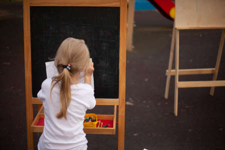 The child draws at the easel on the street. Painting lessons in the open air. A young artist is painting a picture. A girl in a white T-shirt sits with her back.の写真素材