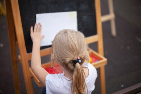 The child draws at the easel on the street. Painting lessons in the open air. A young artist is...