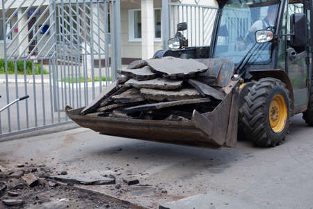 Broken asphalt is removed with a truck bucket. Road repair in the city. Removing the road surface. The worker controls the machinery. Pieces of asphalt.の写真素材