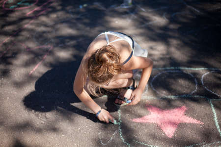 The child draws with chalk. The girl on the asphalt creates a drawing. Summer walk with a child. Entertainment in the yard. Girl artist.の写真素材