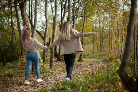 The girls hold hands and walk through the forest. Girlfriends on a walk in the park in autumn. The girls show joy. Two young women togetherの写真素材