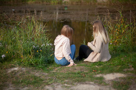 The girls are sitting on the shore of the lake. Women are resting in nature. Scheme from the back. Girls enjoy warm weather in autumn.の写真素材