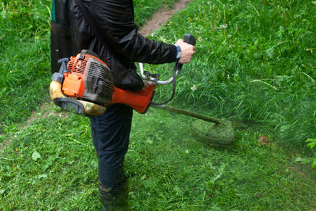 Mows the grass with a hand-held lawn mower. The gardener is cutting the lawn. The gardener's tool cuts tall grass. The lawn mower in action.の写真素材