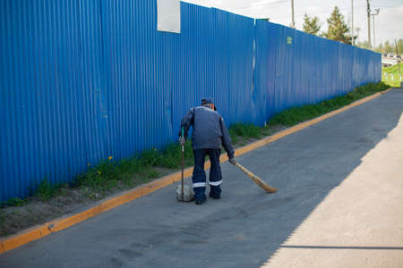 Road cleaner. The janitor sweeps the area with a broom. Removing garbage from the road. Putting things in order near the fence. Cleaning in the parking lot. The cleaner is working.の写真素材