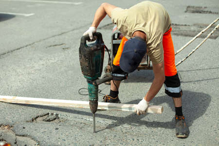 He removes the asphalt with a jackhammer. Removing the asphalt layer from the road. The worker holds a jackhammer. Road repair. Using an electric hammer.の写真素材