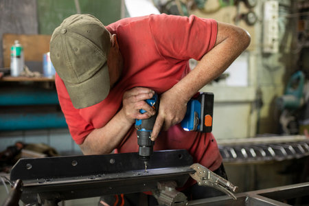 A man drills holes in the metal. The guy works in the garage. Hard man's work. Drilling holes a blank.の写真素材