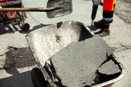 Construction waste trolley. Puts the asphalt into the cart with a shovel. A worker is laying asphalt. Road repair. Destruction of old asphalt.の写真素材