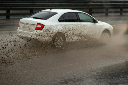 The car is driving through a puddle. Splashes from a puddle on the road. The car, dirty from...