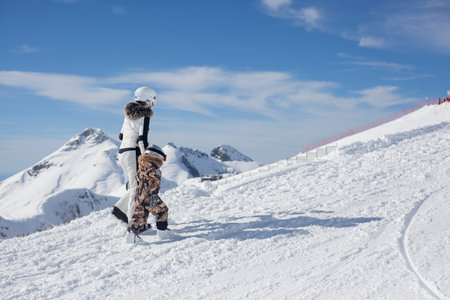 Mom with a child in a ski resort. The family walks in the snow in special ski equipment. Rest high on a snowy slope. A girl leads a child to learn to ride from the mountain in the snow. Frosty sunny day in the mountains.の写真素材