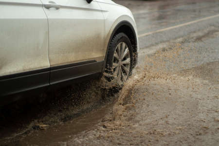 The car is driving in a puddle. White car in the mud. Roads for transport are flooded with water. Street flooding on the highway. Transport and splashes from under the wheels of the car.の写真素材