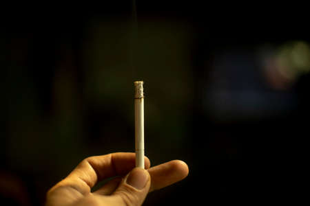 Cigarette in hand on a black background. Smoking is harmful to your health. The tobacco product is pinched in your fingers. The smoker is holding one cigarette.の写真素材