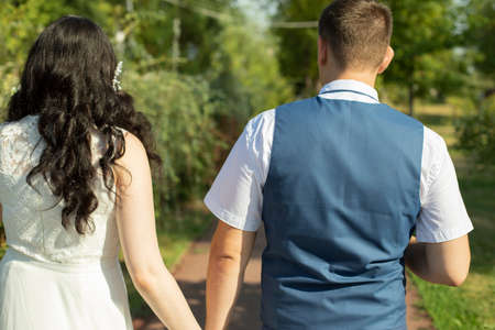 The newlyweds walk along the park path holding hands. A guy and a girl are taking a romantic walk. Lovers walk in the park in summer.の写真素材