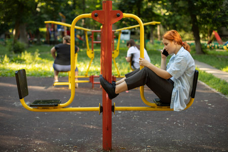 The teenager sits on the playground. The girl speaks on the phone at the playground. Schoolgirl is talking on a smartphone. The girl is sitting alone on a sports simulator.の写真素材