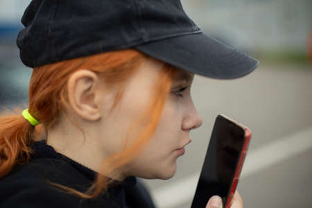The girl records a voice message on the street. The woman communicates on a smartphone. The teenager speaks into the phone screen. Woman in a cap.の写真素材