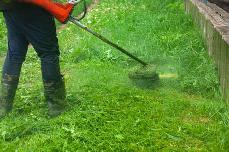 Mows the grass with a hand-held lawn mower. The gardener is cutting the lawn. The gardener's tool cuts tall grass. The lawn mower in action.の写真素材