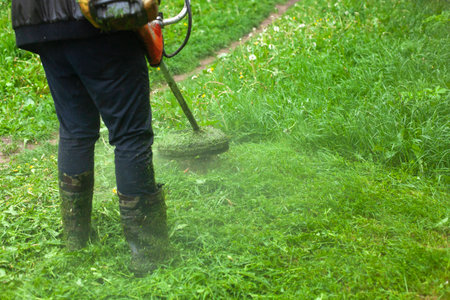 Mows the grass with a hand-held lawn mower. The gardener is cutting the lawn. The gardener's tool cuts tall grass. The lawn mower in action.の写真素材