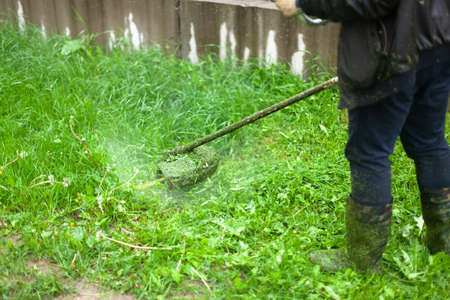Mows the grass with a hand-held lawn mower. The gardener is cutting the lawn. The gardener's tool cuts tall grass. The lawn mower in action.の写真素材