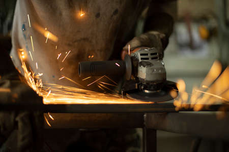 Metal processing with a grinder. Sparks from the tool. A man in the workshop wearing gloves with a grinder in his hand. Sparks from grinding steel.の写真素材
