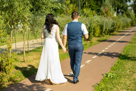 The newlyweds walk along the park path holding hands. A guy and a girl are taking a romantic walk. Lovers walk in the park in summer.の写真素材