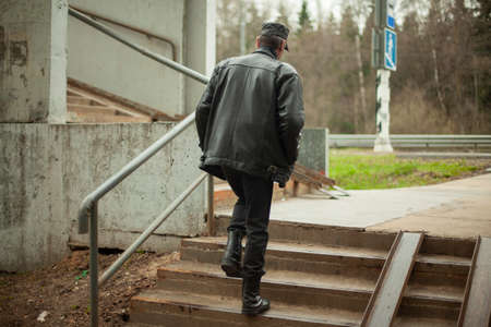 The man climbs the stairs. A person walks up the steps. Old pedestrian crossing. Place of crossing the road. A man in black leather clothes. Leather jacket and cap on an old man in Russia.の写真素材