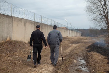 Men with spades are walking along the road. Workers walk to dig a trench. The guys go to work. Men walk along the fence. Shooting from the back.の写真素材