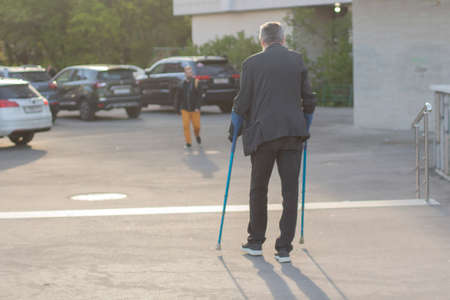 A man with crutches in the city. The guy broke his leg. Difficulty of movement of a disabled person around the city. Damaged limb. A man walks in the square.の写真素材