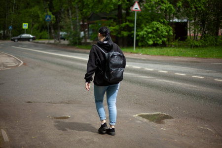 The girl walks with a backpack. A woman walks along the footpath. Photo of a woman from the back.