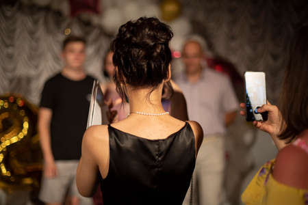 A girl takes pictures of the family on a smartphone. A schoolgirl in a black dress takes off on her phone. View from the back. A woman takes a memorable photo.の写真素材