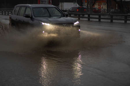 The car is driving in a puddle. Sprays from under the wheels. A flooded highway. Transport with lights turned on goes on the highway.の写真素材