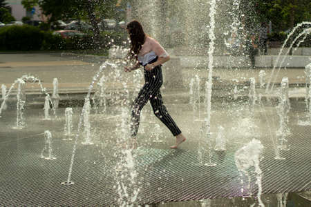 A child runs among the jets of water in the town square. Splashes of the fountain in the summer. A hot day in the city and a source of water.の写真素材