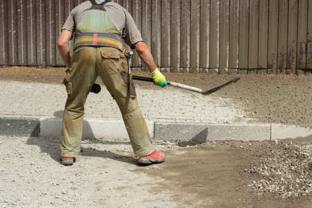 Work with a shovel at a construction site. A worker throws cement and gravel with a shovel. Cleaning the road from scattered building material.の写真素材