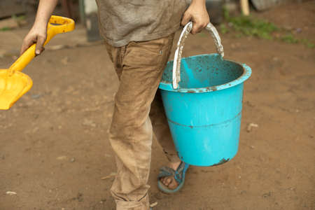 The child is holding a bucket. Blue bucket in hand. The boy plays with a bucket and a shovel. A child stained his clothes while playing in the yard.の写真素材