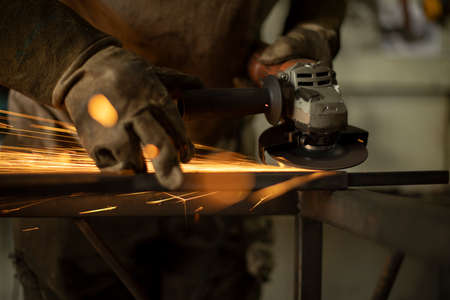Metal processing with a grinder. Sparks from the tool. A man in the workshop wearing gloves with a grinder in his hand. Sparks from grinding steel.の写真素材