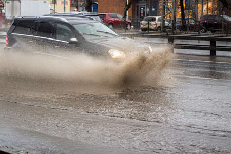 The car is driving in a puddle. Sprays from under the wheels. A flooded highway. Transport with lights turned on goes on the highway.の写真素材