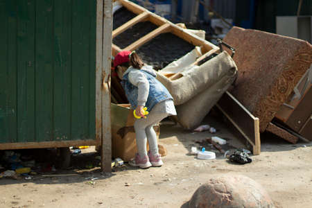 A child in a junkyard. The girl is dripping in the trash. The child found the toy in the trash heap. The poor girl is walking alone.の写真素材