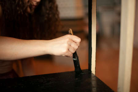 A girl paints furniture at home. Painting with a brush in the black color of a wooden surface. Brush in the girl's hand.の写真素材