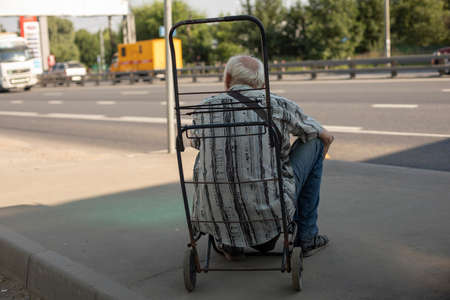 An old man at a bus stop. Grandpa is sitting on a cart. The pensioner is waiting for transport. The gray-haired man is tired. A man in the shade from the heat.の写真素材