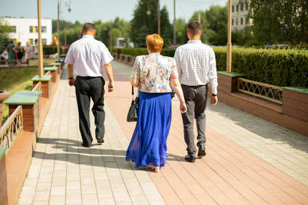 People are walking down the street of the city, the view from behind. Three adults on a walk, men in shirts and trousers, a woman in a blue skirt. On the sides a lot of greenery, a sunny day in the cityの写真素材