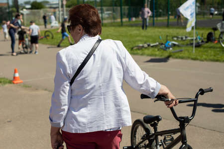 An elderly woman in a white jacket with a bicycle. Sunny day, city park. Competitions. City Lifestyleの写真素材