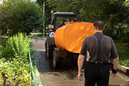A man walks the street behind a tractor. A man in trousers with suspenders and a T-shirt, an orange cistern, a sunny day. Technique used to clean the streets. Urban lifeの写真素材