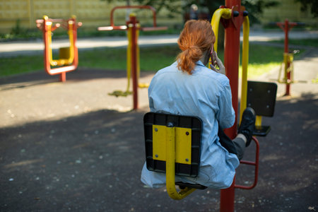 A girl with red hair in a denim shirt is talking on the phone at the playground. Solo day, rest after school. City Lifestyleの写真素材