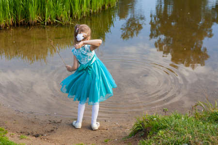 A girl in a blue dress throws pebbles into the water. A child plays with the river in nature. A smartly dressed baby is having fun near the lake. Childhood without the Internet. Healthy lifestyle baby.の写真素材