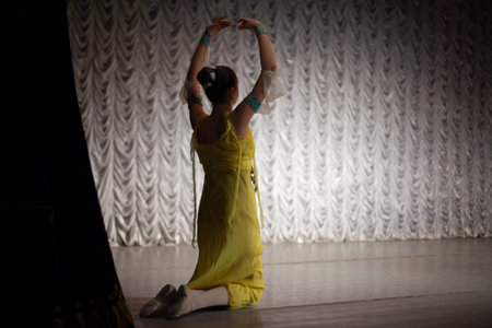 The girl sits on her knees and raised her hands up. Girl in a yellow dress. Dancer's movements in the assembly hall. Artificial lighting on the stage.の写真素材